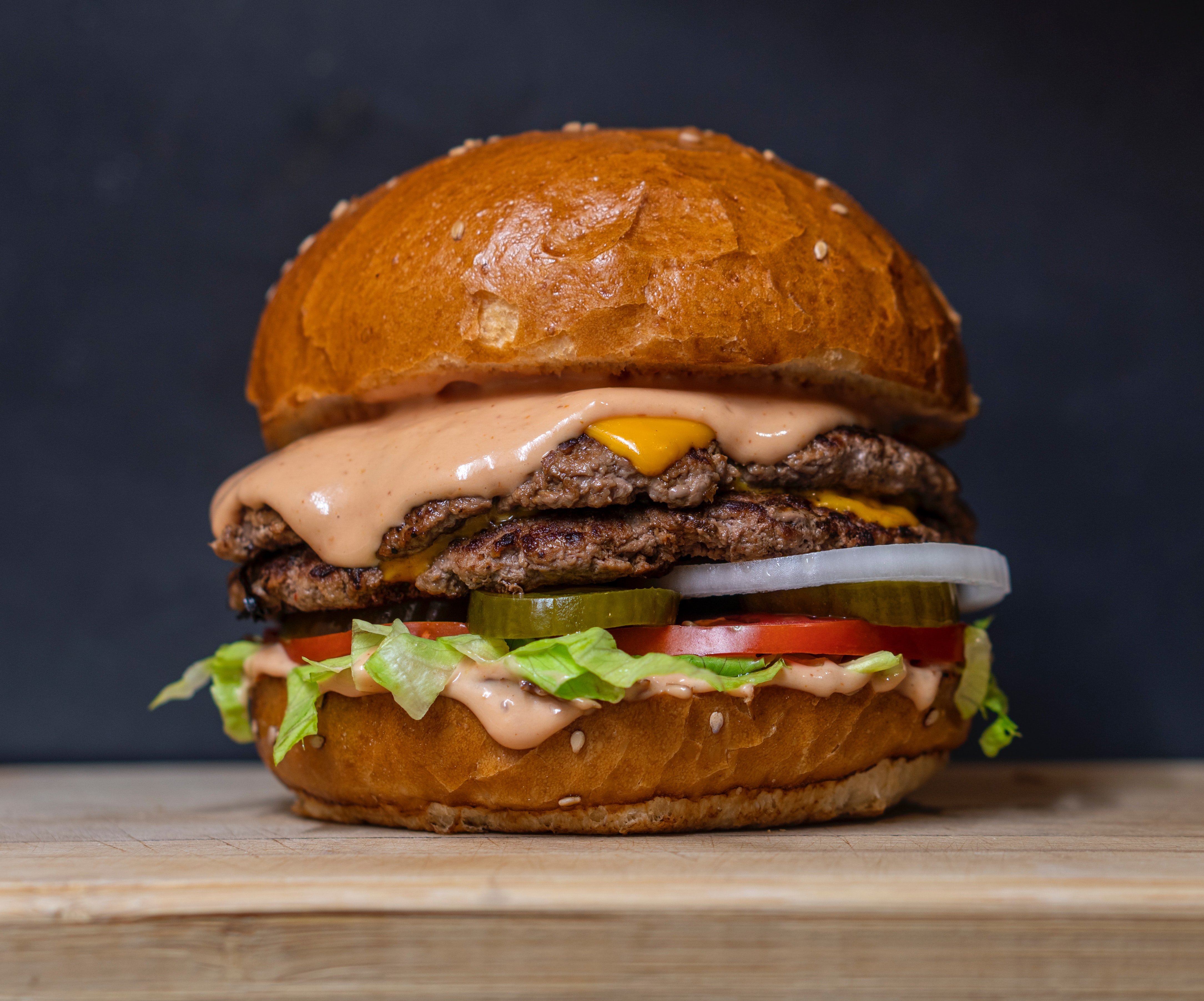 A close up of a burger with the following ingredients in order from top to bottom: a sesame seed bun, sauce, 
              cheese, two patties, pickles, onions, tomato, lettuce, and more sauce, bottom bun on a clean wood table.
