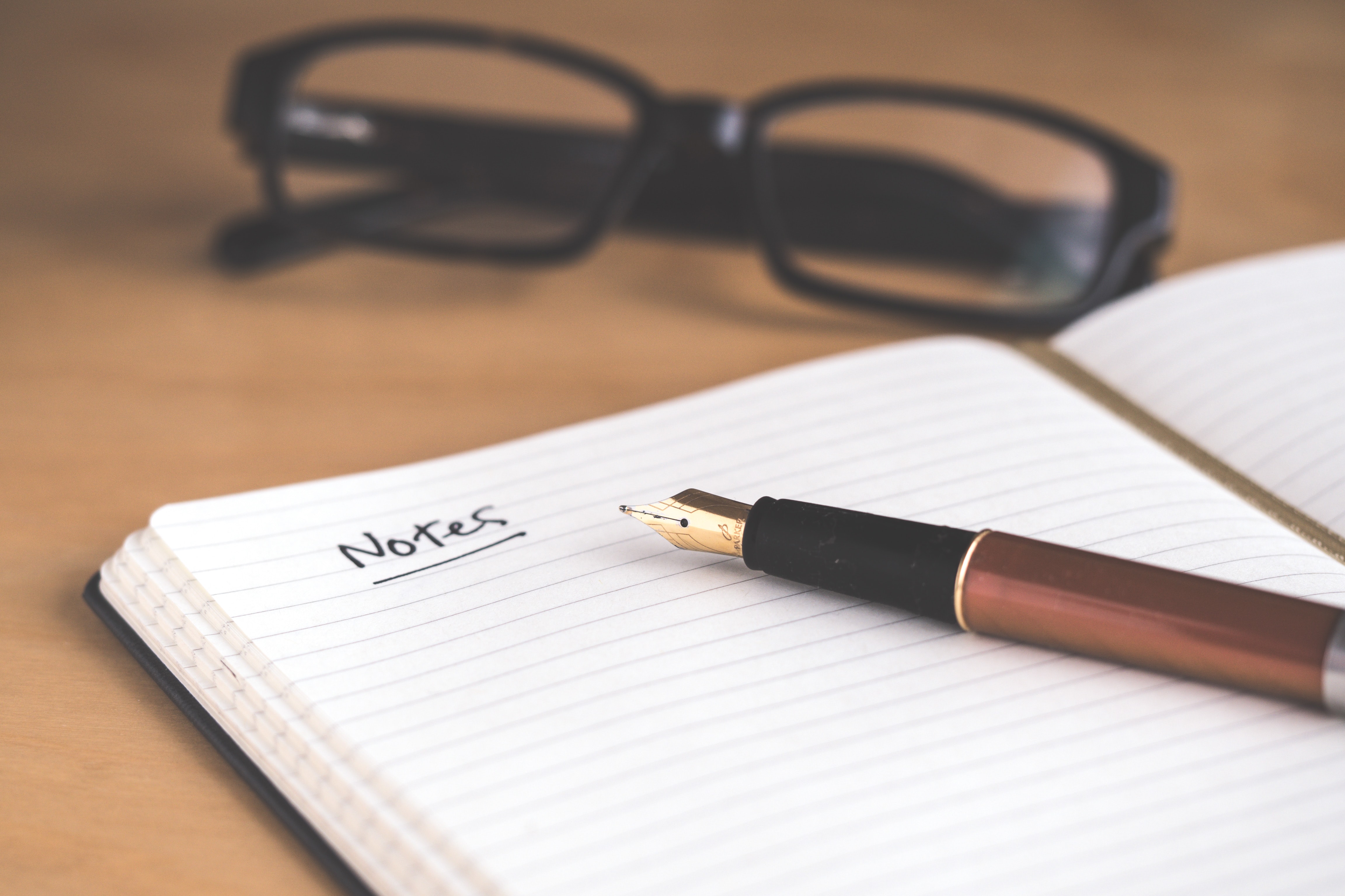 A table with a pair of glasses with short lenses and black 
              frames resting above an open notebook with a pen laying on it and the word 'notes' written at the top of the page